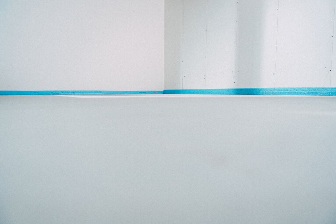 Ein weißer Raum mit glatten Wänden und einem glatten, frisch gegossenen Boden. Blaue UZIN Randdämmstreifen säumt den unteren Teil der Wände, was auf kürzliche Bau- oder Renovierungsarbeiten hindeutet.
