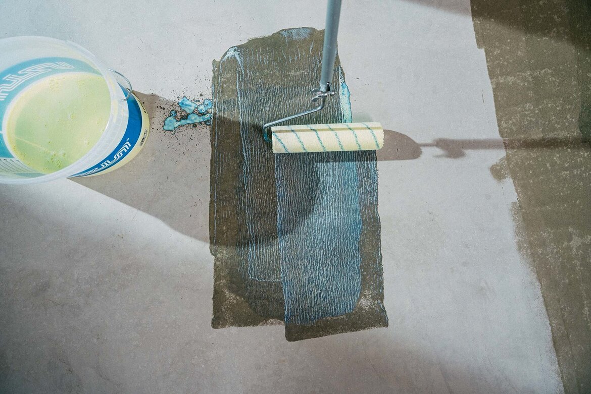 Ein Farbroller trägt eine flüssige Grundierung UZIN Hydro Primer 1 auf einen Betonboden auf, wobei ein Eimer mit der Flüssigkeit in der Nähe steht. Durch die Grundierung entsteht ein dunkler, nass aussehender rechteckiger Streifen auf der ansonsten trockenen Oberfläche.