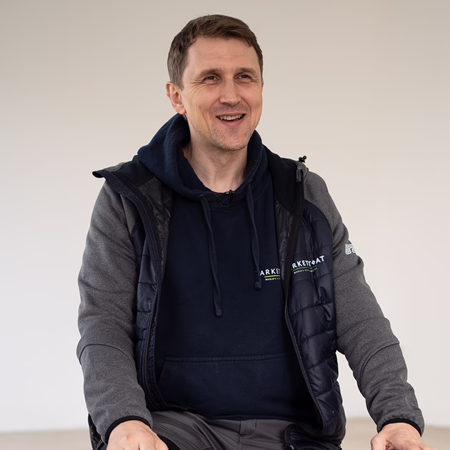 Ein Mann sitzt lächelnd da und trägt einen dunkelblauen Kapuzenpulli und eine graue Jacke. Der Hintergrund ist einfarbig und hell.
