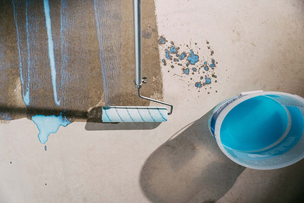Ein Farbroller trägt leuchtend blaue Grundierung UZIN PE 360 PLUS auf, in der Nähe befinden sich blaue Farbspritzer. Ein mit blauer Dispersionsgrundierung gefüllter Eimer steht auf einem hellen Boden und wirft einen Schatten.
