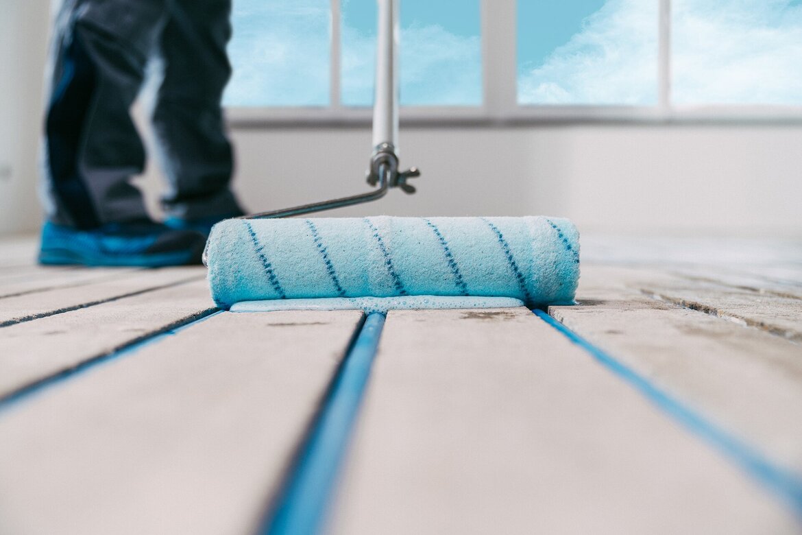 Nahaufnahme einer Nylonplüschrolle, die einen hellblauen Anstrich, Grundierung UZIN PE 360 Plus, aufträgt. Im Hintergrund sind die Beine und blauen Schuhe einer Person zu sehen, die vor einem hellen Fenster mit blauem Himmel steht.