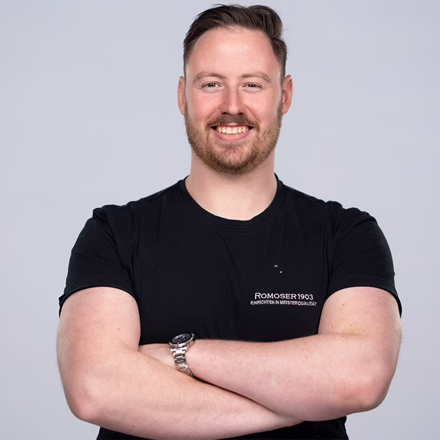 Ein lächelnder Mann mit kurzen Haaren, Bart und schwarzem T-Shirt steht mit verschränkten Armen vor einem schlichten hellgrauen Hintergrund. Er trägt eine Uhr am linken Handgelenk.