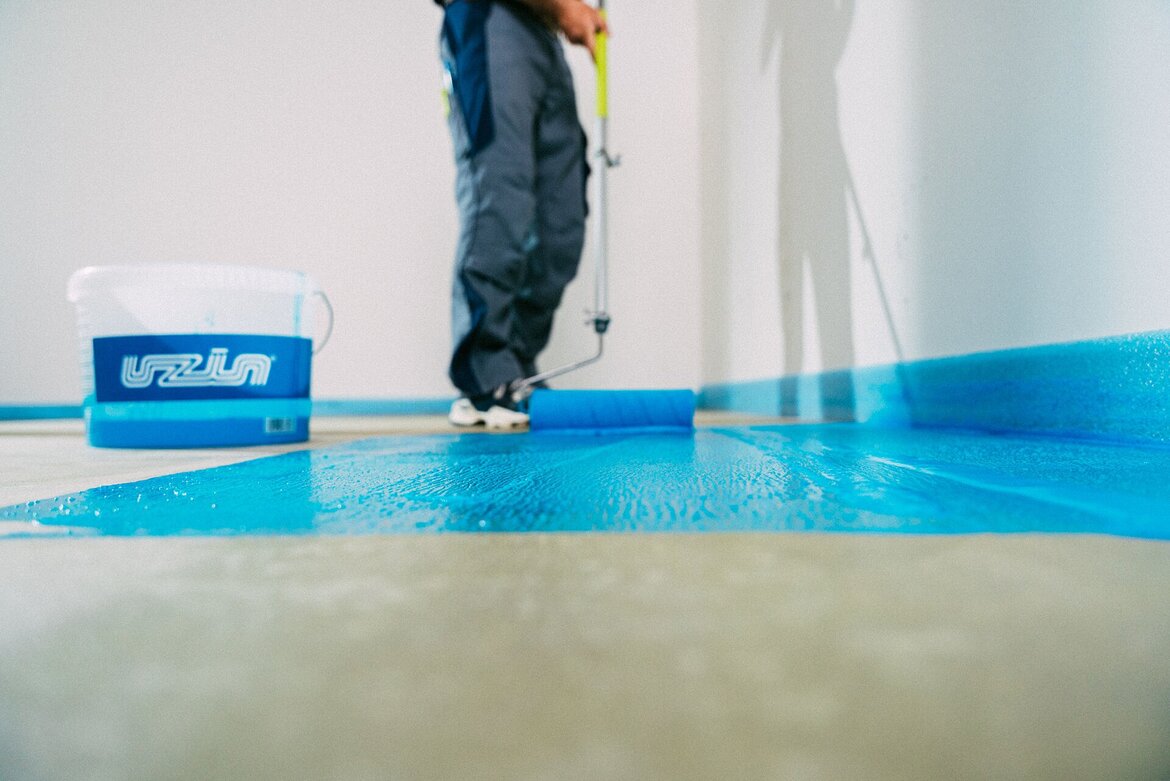 Eine Person trägt mit einer Farbrolle hellblaue Grundierung, UZIN Hydro Primer 2, auf den Boden in der Nähe einer Wand auf. Ein Eimer mit der Aufschrift Uzin steht daneben, und das Gesicht der Person ist auf dem Bild nicht zu sehen.