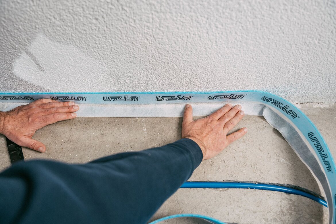 Zwei Hände drücken einen blau-weißen Klebestreifen, den UZIN Randdämmstreifen mit der Aufschrift UZIN auf die Kante, wo ein Betonboden auf eine strukturierte weiße Wand trifft. In der Nähe sind blaue Schläuche der Fussbodenheizung auf dem Boden zu sehen.
