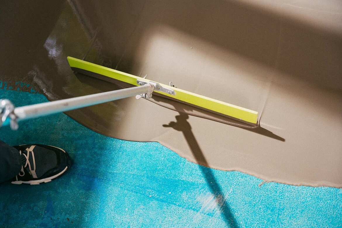 Eine Person verteilt mit einer WOLFF Rakel graue, flüssige Bodenausgleichsmasse, UZIN Hydro LevelStar, auf einer blauen Fläche, um den Boden für einen neuen Belag vorzubereiten.