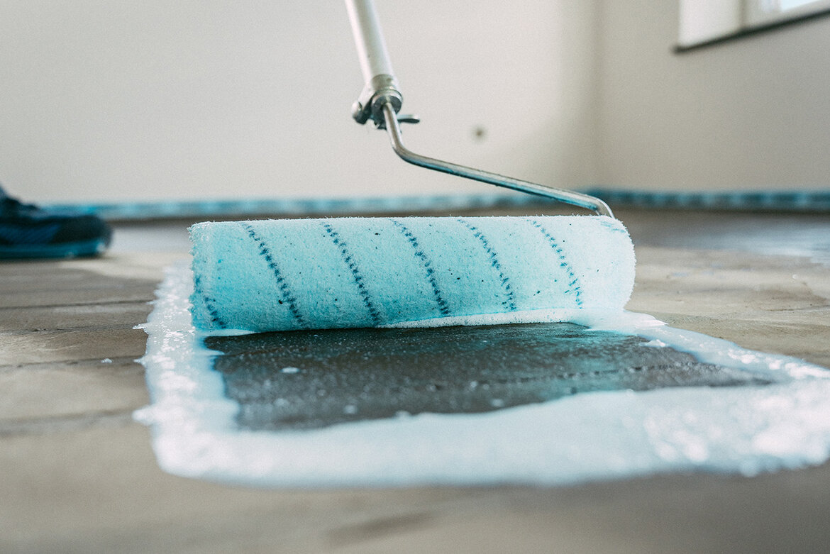 Ein Farbroller trägt eine Schicht hellblaue Dispersions-Grundierung UZIN PE 360 Plus auf einen Betonboden in einem Raum mit hellen Wänden und einem Fenster auf, während Randdämmstreifen die Ränder säumen.