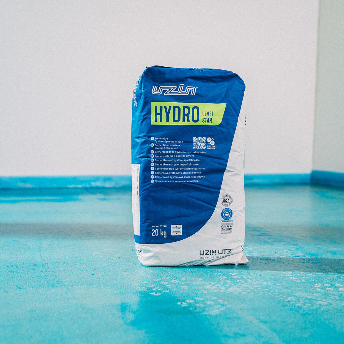 Ein 20-kg-Sack UZIN Hydro Level Star steht auf einem blau grundierten Untergrund vor einer weißen Wand. Der Sack ist überwiegend blau und weiß, mit Produktinformationen auf der Vorderseite.